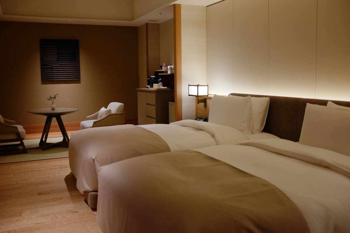 A hotel room with two beds, a desk, a chair, a small table with a vase, and a wall-mounted lamp.