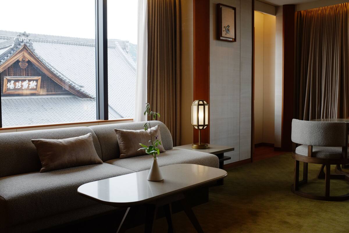 A modern hotel room with a sofa, a small table, a chair, a lamp, and a view of traditional architecture outside the window.