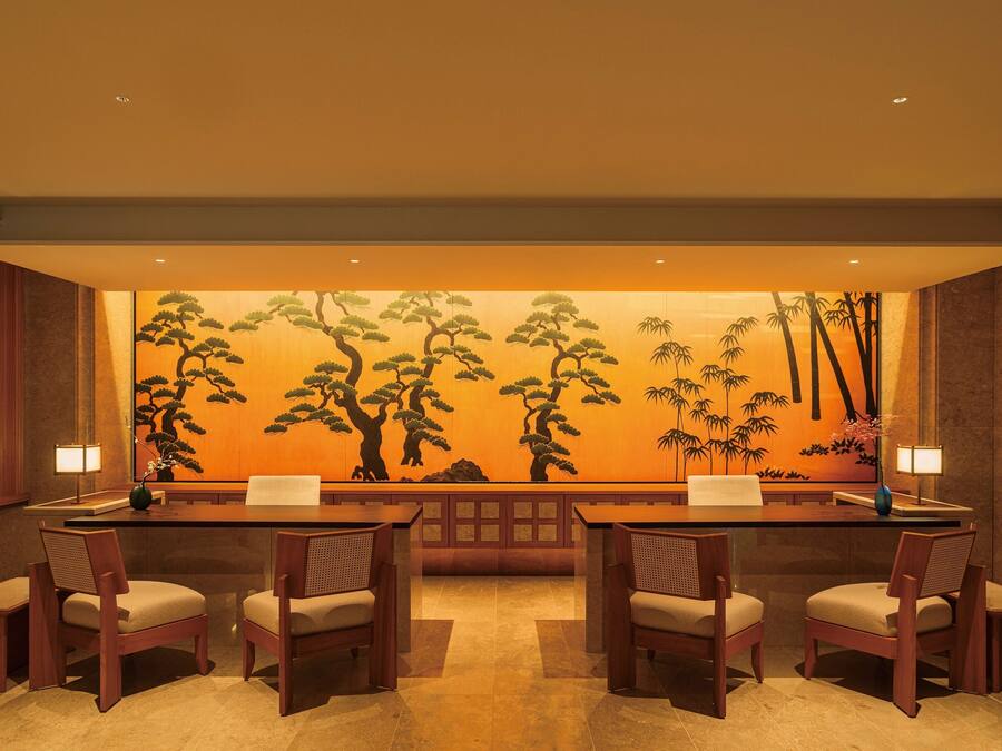 A traditional Japanese-style room with a long table and four chairs, a large wall mural of trees, and two lamps on the table.
