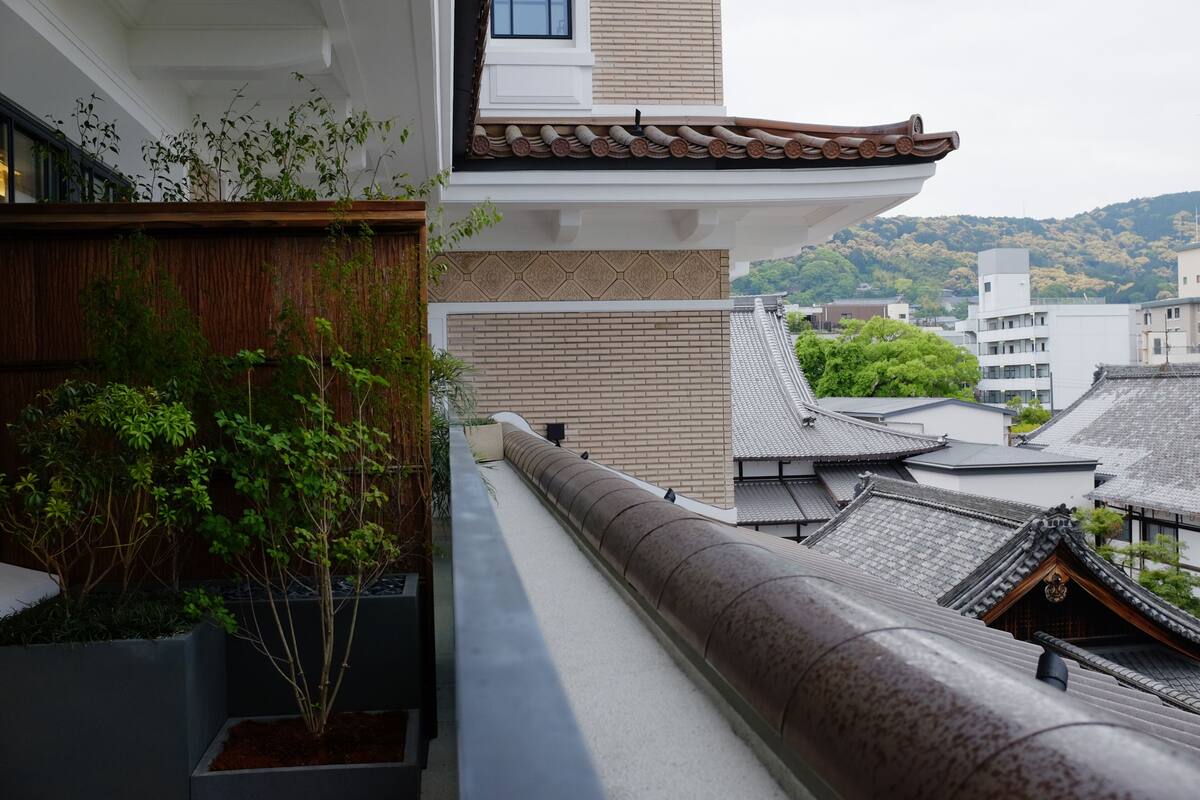 A traditional Japanese building with a sloped roof, a wooden fence, and a view of other buildings and hills in the background.