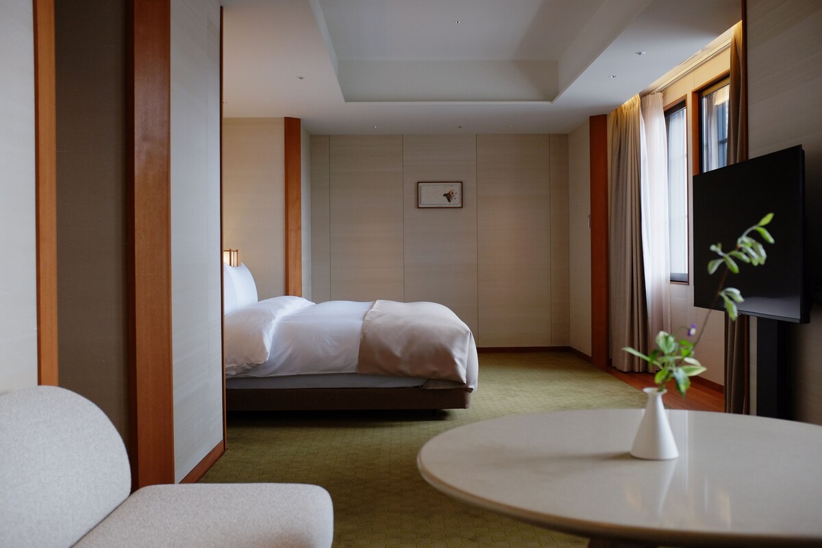 A hotel room with a bed, a TV, a small table with a vase, and a chair.