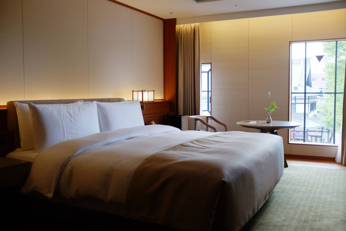 A hotel room with a large bed, a small table, and a window with a view.