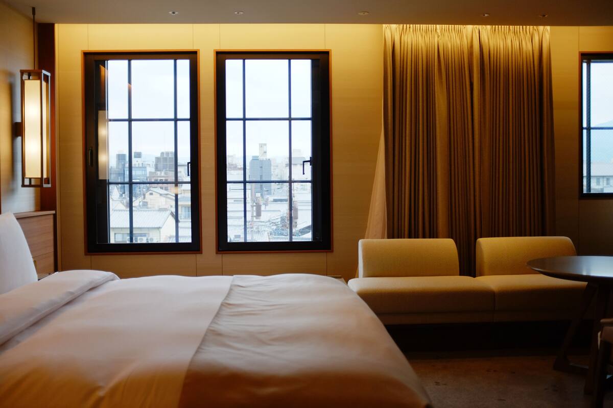 A hotel room with a large bed, two sofas, a small table, and two windows offering a city view.