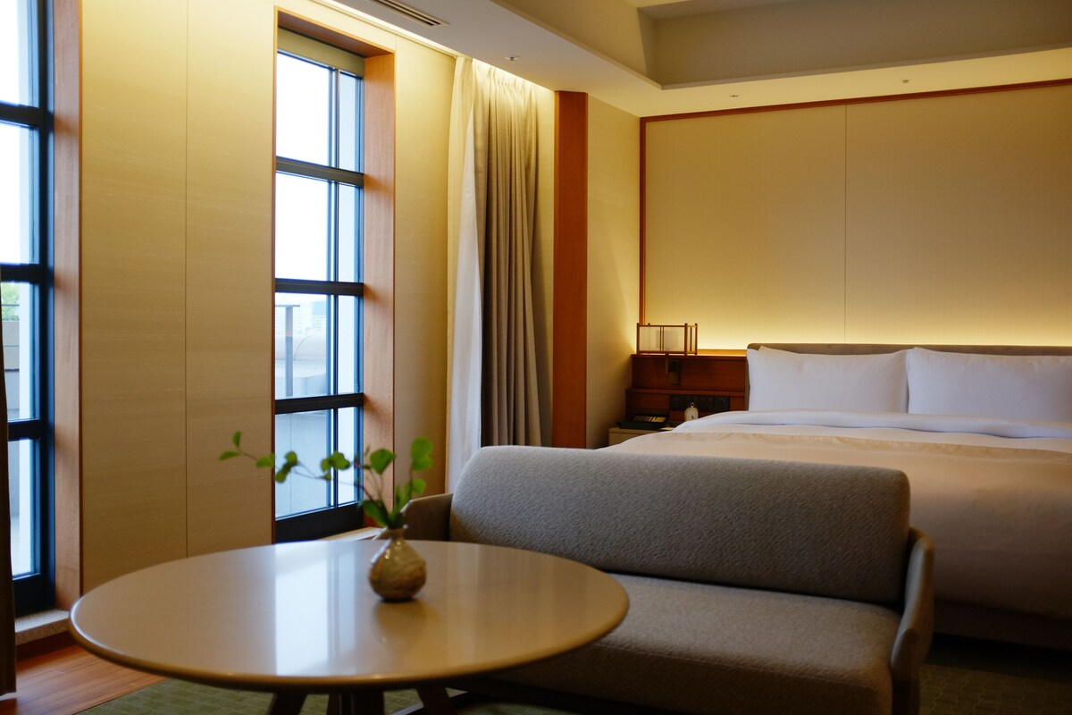 A hotel room with a bed, a sofa, a table with a vase, a nightstand, and a window with curtains.