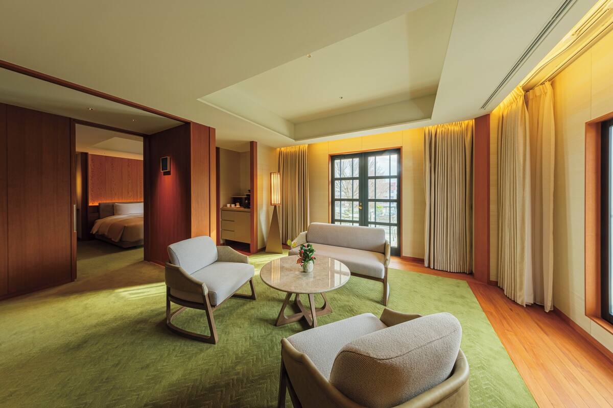 A hotel room with a green carpet, two armchairs, a sofa, a round coffee table, and a bed in the background.
