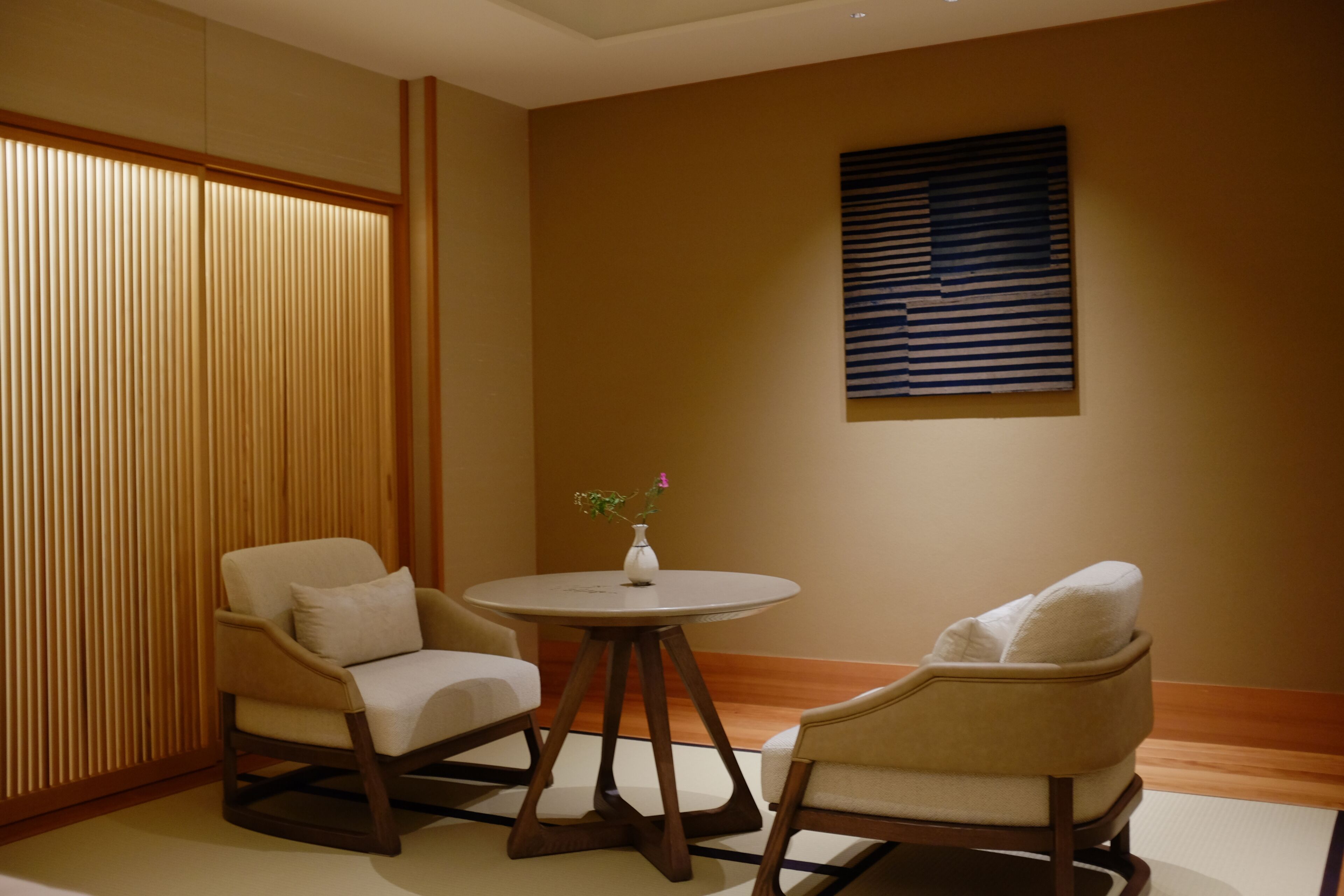 A room with two chairs, a small table, a vase with flowers, a sliding door, and a wall-mounted artwork.