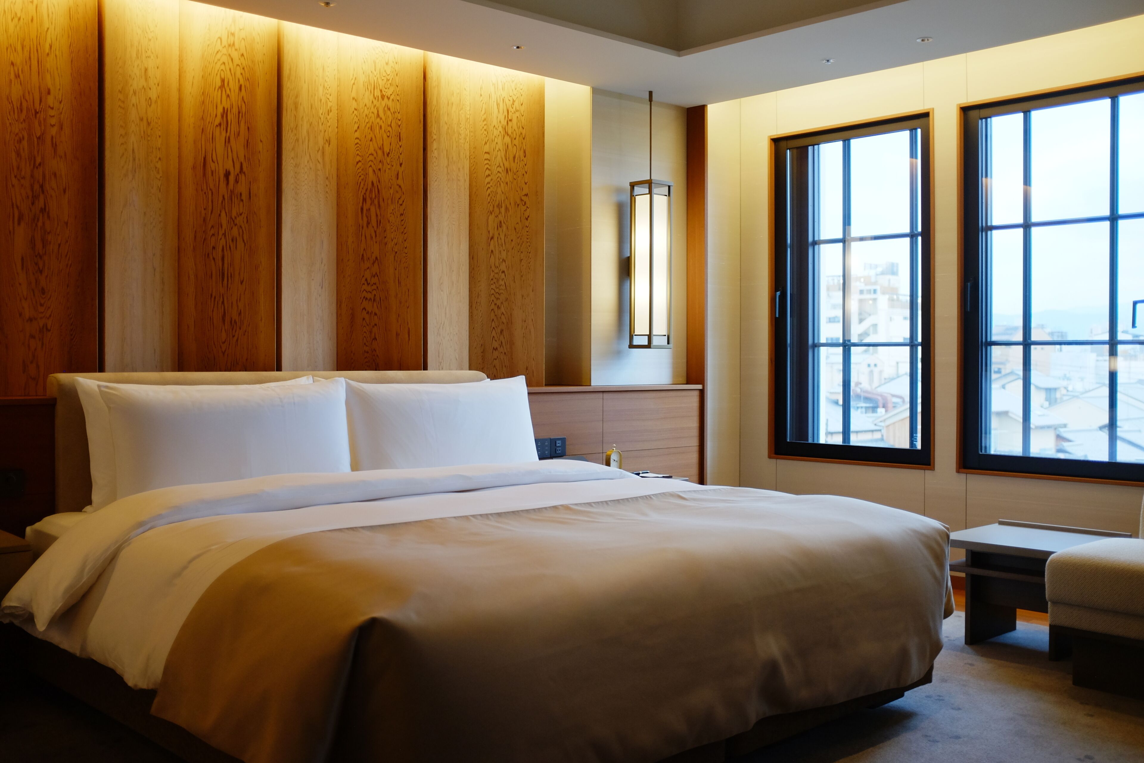 Habitación de hotel con una cama grande, pared de paneles de madera, una mesita de noche, un escritorio y una ventana con vista a la ciudad.