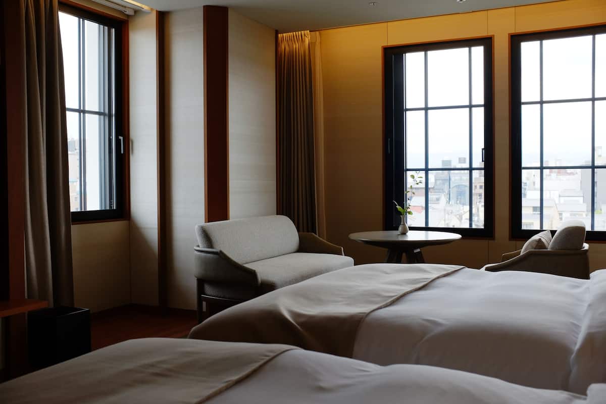 A hotel room with two beds, a chair, a small table, and large windows with a city view.