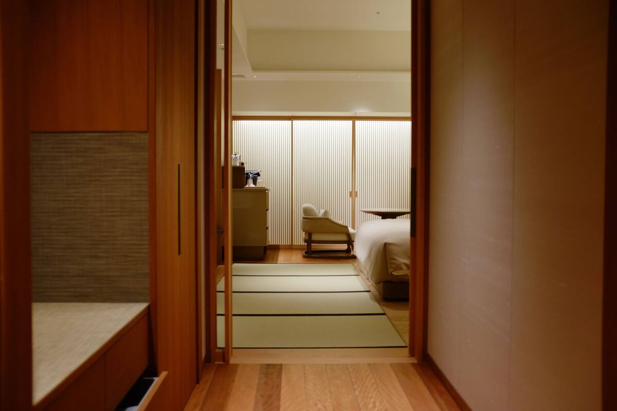 A narrow hallway leading to a room with a bed, a chair, and a small table.