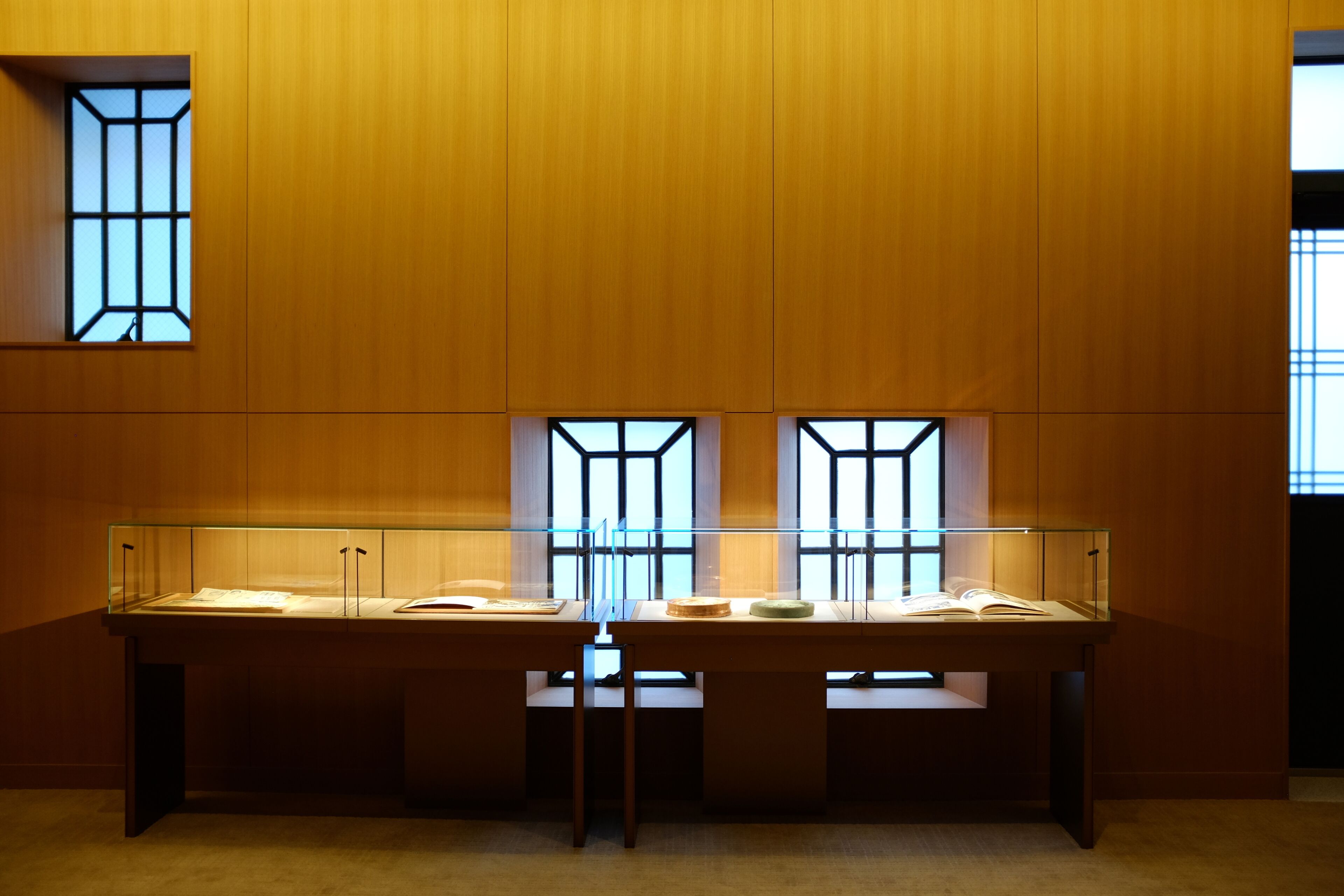 A room with wooden panel walls, a glass display case with books, and a window with a geometric design.