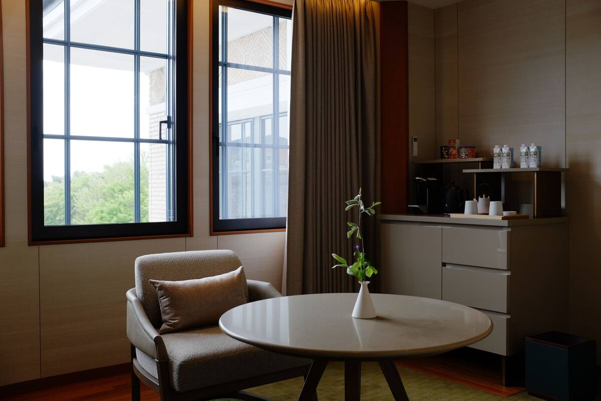 A modern room with a sofa, a round table, a window with curtains, and a kitchenette with cabinets and appliances.