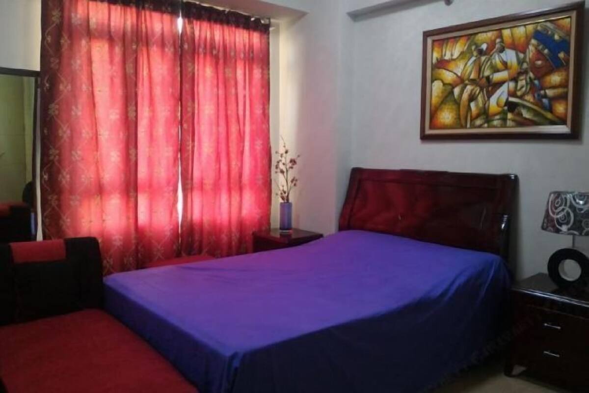 A bedroom with a bed, a nightstand, a lamp, a painting on the wall, and a window with curtains.