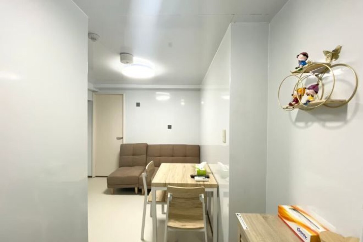 A small, well-lit room with a wooden dining table, two chairs, a brown sofa, and a wall-mounted shelf with decorative items.