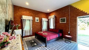 Quarto com parede de tijolos, cama com colcha vermelha, mesa de cabeceira com um vaso de flores e janela com cortinas.