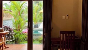 Une pièce avec une chaise en bois, une petite table et une vue sur un jardin agrémenté de plantes et d’une piscine.