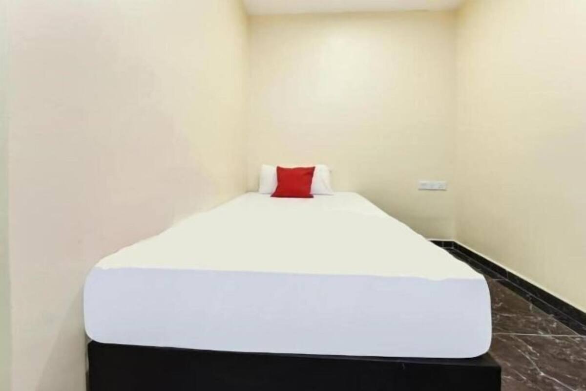 A narrow hotel room with a single bed, a red pillow, and a black headboard.