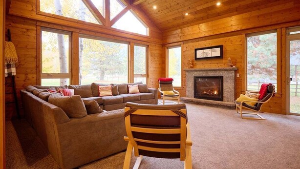 A log cabin interior with a sectional sofa, a fireplace, and large windows. - Mountain Lodge w/ Hot Tub + Private River Beach (Leavenworth)