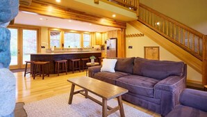 A spacious living area with a wooden staircase, a sofa, a coffee table, and a kitchen in the background. - Mountain Lodge w/ Hot Tub + Private River Beach (Leavenworth)