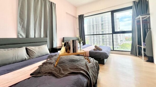 A modern bedroom with a large window, a bed with a wooden headboard, a bedside table with books and a vase, and a small seating area with a cushion. - Sunway Geo Avenue Next to Medical Center Sunway University and Taylor University (Subang Jaya)