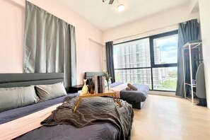 A modern bedroom with a large window, a bed with a wooden headboard, a bedside table with books and a vase, and a small seating area with a cushion. - Sunway Geo Avenue Next to Medical Center Sunway University and Taylor University (Subang Jaya)