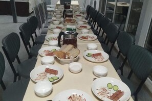 Mesa longa posta com pratos, tigelas e uma cafeteira, rodeada por cadeiras.