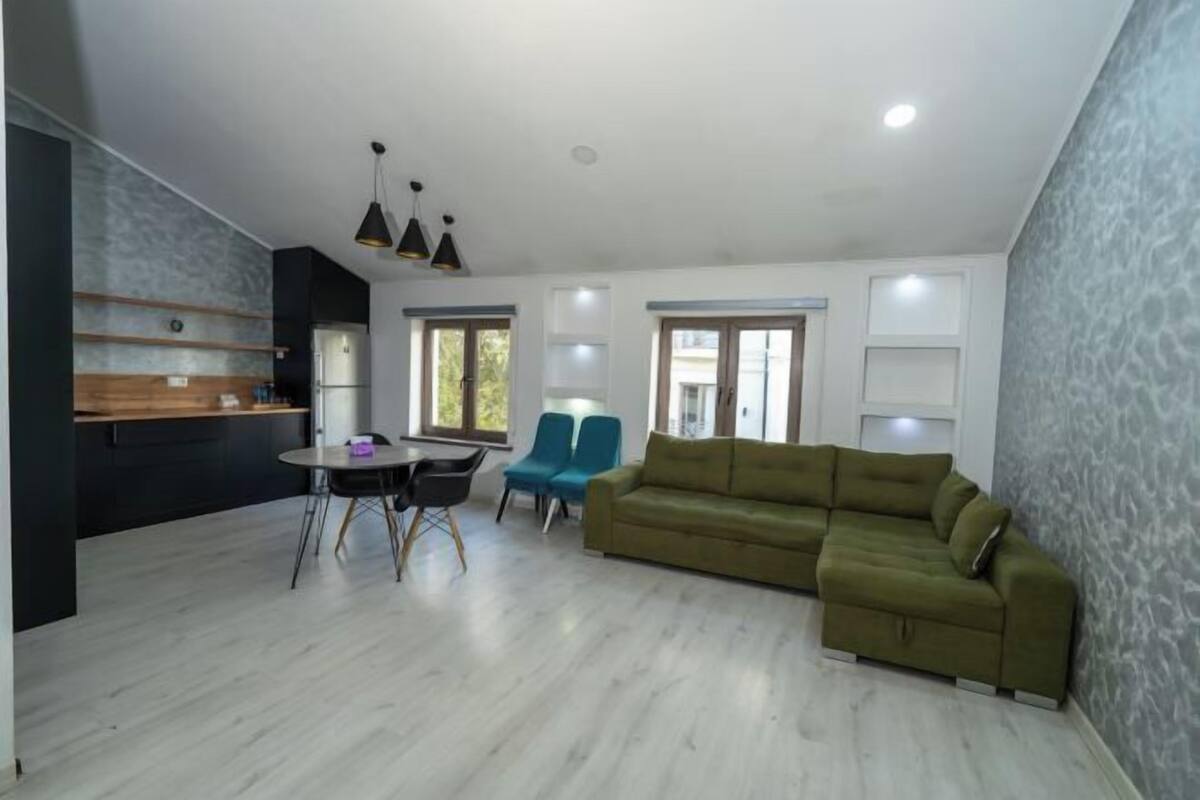 Ein modernes Wohnzimmer mit einem grünen Sofa, einem Esstisch mit Stühlen und einer Küchenzeile mit Kühlschrank und Holzregalen.