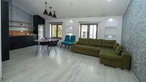 A modern living room with a green sofa, a dining table with chairs, and a kitchen area with a refrigerator and wooden shelves. - Central Asia Inn Hotel (Almaty)