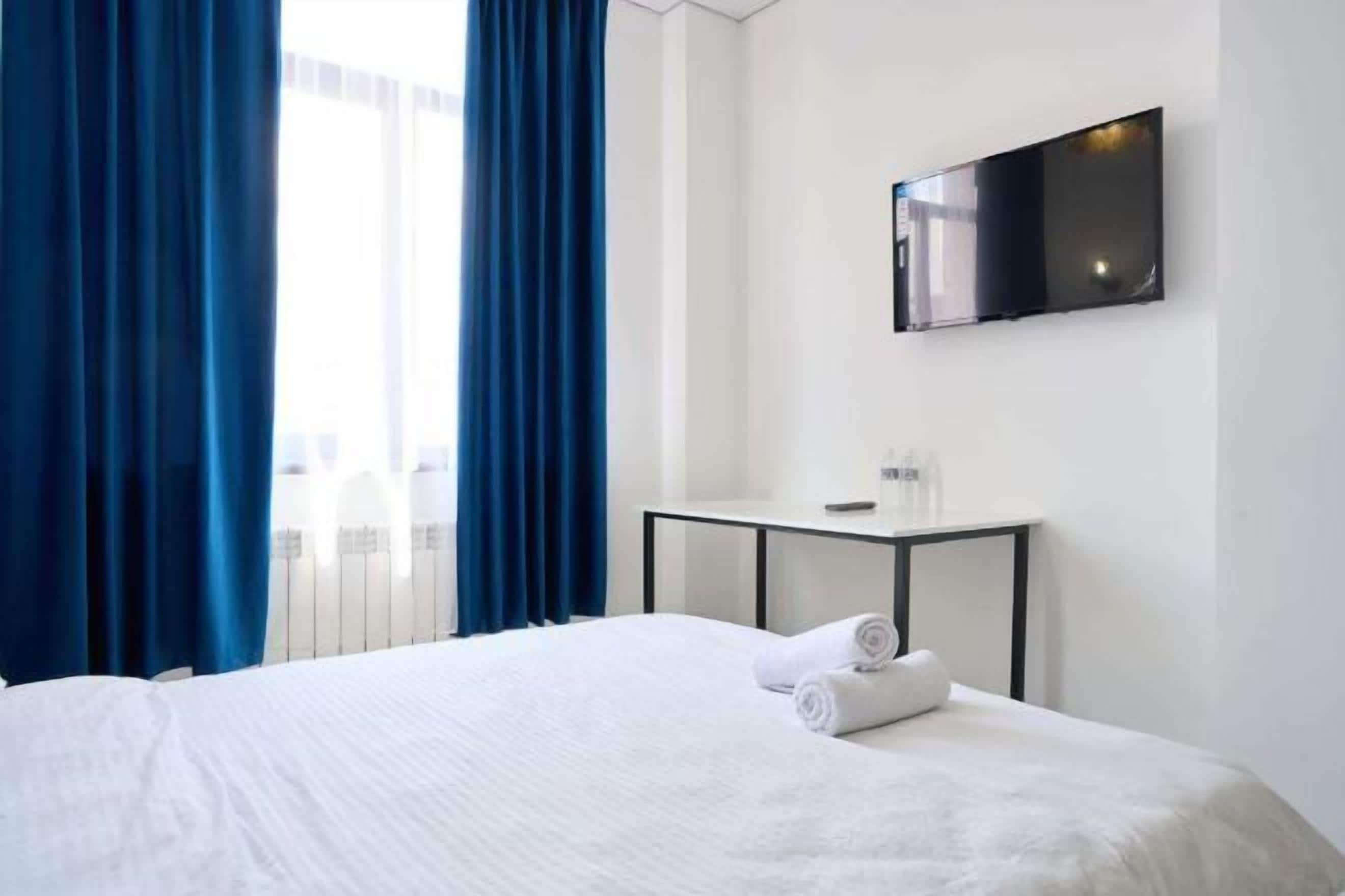 A hotel room with a bed, a flat-screen TV mounted on the wall, a small table with a remote control, and blue curtains.