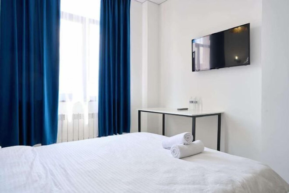 A hotel room with a bed, a flat-screen TV mounted on the wall, a small table with a remote control, and blue curtains.