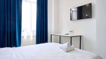 A hotel room with a bed, a flat-screen TV mounted on the wall, a small table with a remote control, and blue curtains.