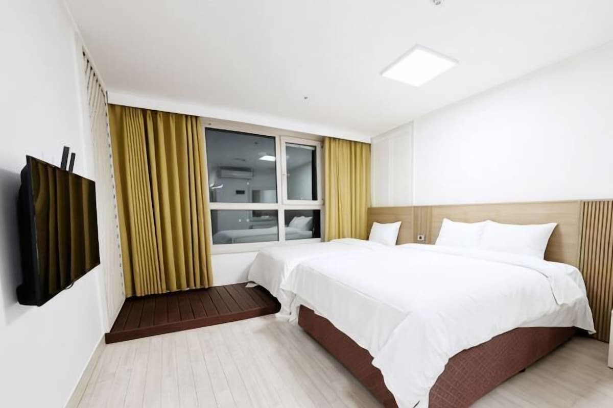 A hotel room with a bed, a TV, a window with curtains, and a wooden headboard.