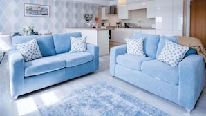A modern living room with two sofas, a kitchen in the background, and a patterned rug. - Harbour View - 2 Bedroom Apartment - Milford Haven (Milford Haven)