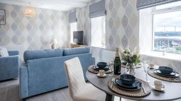 A modern living room with a sofa, dining table set with plates, glasses, and a bottle, and a TV on a stand.