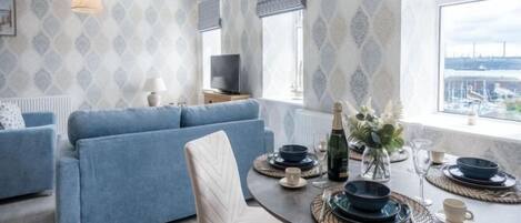 A modern living room with a sofa, dining table set with plates, glasses, and a bottle, and a TV on a stand.