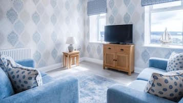 A room with a patterned wallpaper, a television on a wooden cabinet, and a view of the sea through a window.