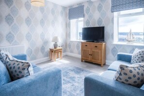 A room with a patterned wallpaper, a television on a wooden cabinet, and a view of the sea through a window. - Harbour View - 2 Bedroom Apartment - Milford Haven (Milford Haven)
