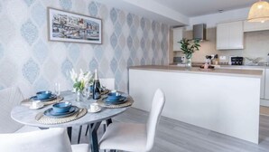 A modern kitchen with a central island, white cabinetry, and a dining area with a round table set for two. - Harbour View - 2 Bedroom Apartment - Milford Haven (Milford Haven)