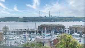 A marina with boats docked, a building with a red facade, and industrial structures in the background. - Harbour View - 2 Bedroom Apartment - Milford Haven (Milford Haven)