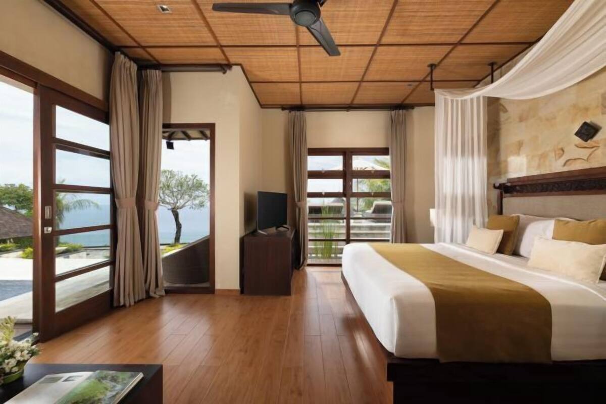 A bedroom with a bed, a ceiling fan, a TV, and large windows.
