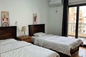 A hotel room with two single beds, a wooden nightstand with a lamp, a wall-mounted air conditioner, and a balcony with a view of buildings. - Qinfu Hotel (Cairo)
