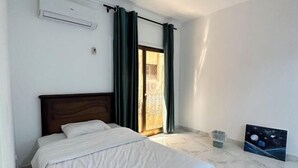 Une chambre à coucher avec un lit, une fenêtre avec des rideaux, un tableau au mur et une poubelle.