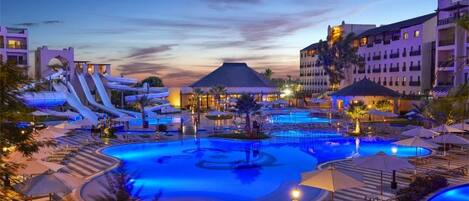 Zona de piscina en un resort con múltiples toboganes y una fuente central, rodeada de edificios y palmeras.