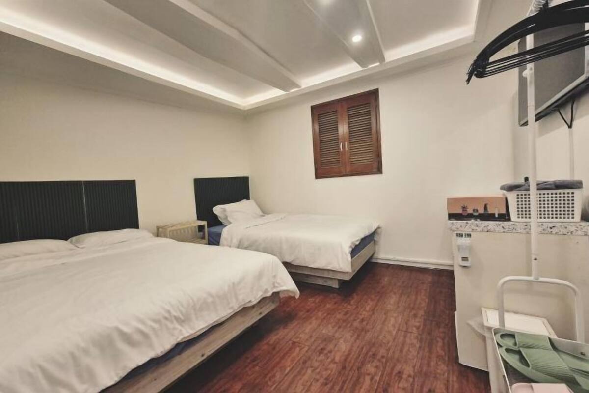 A room with two beds, a small table, a chair, a window with shutters, and a ceiling light fixture.