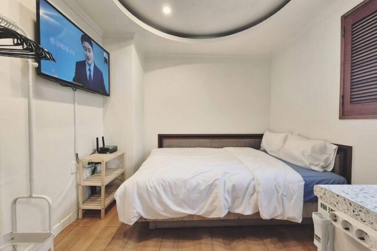 A bedroom with a bed, a TV mounted on the wall, a wooden shelf, and a window with blinds.
