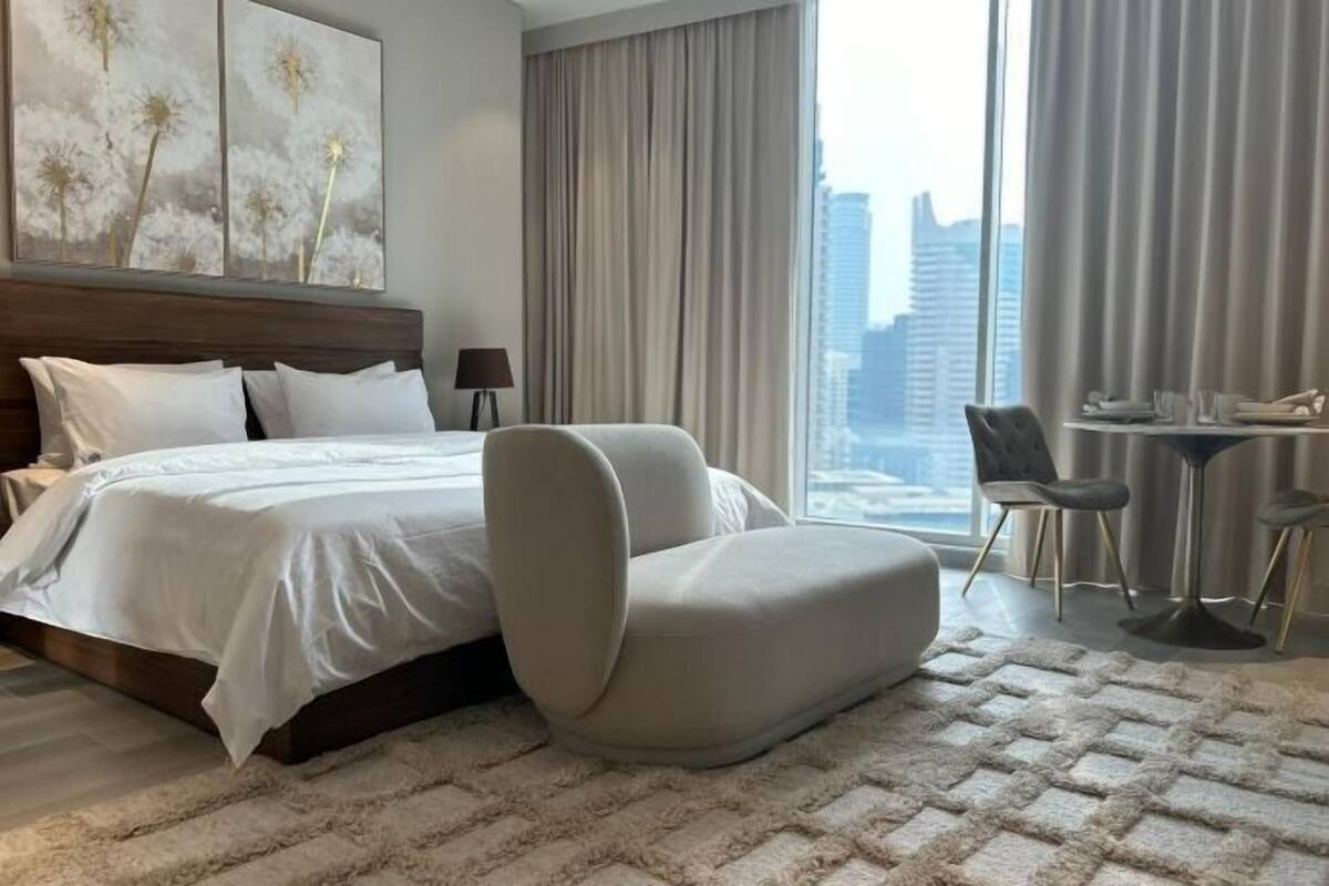 A hotel room with a bed, a sofa, a dining table with chairs, and a large window with a city view.