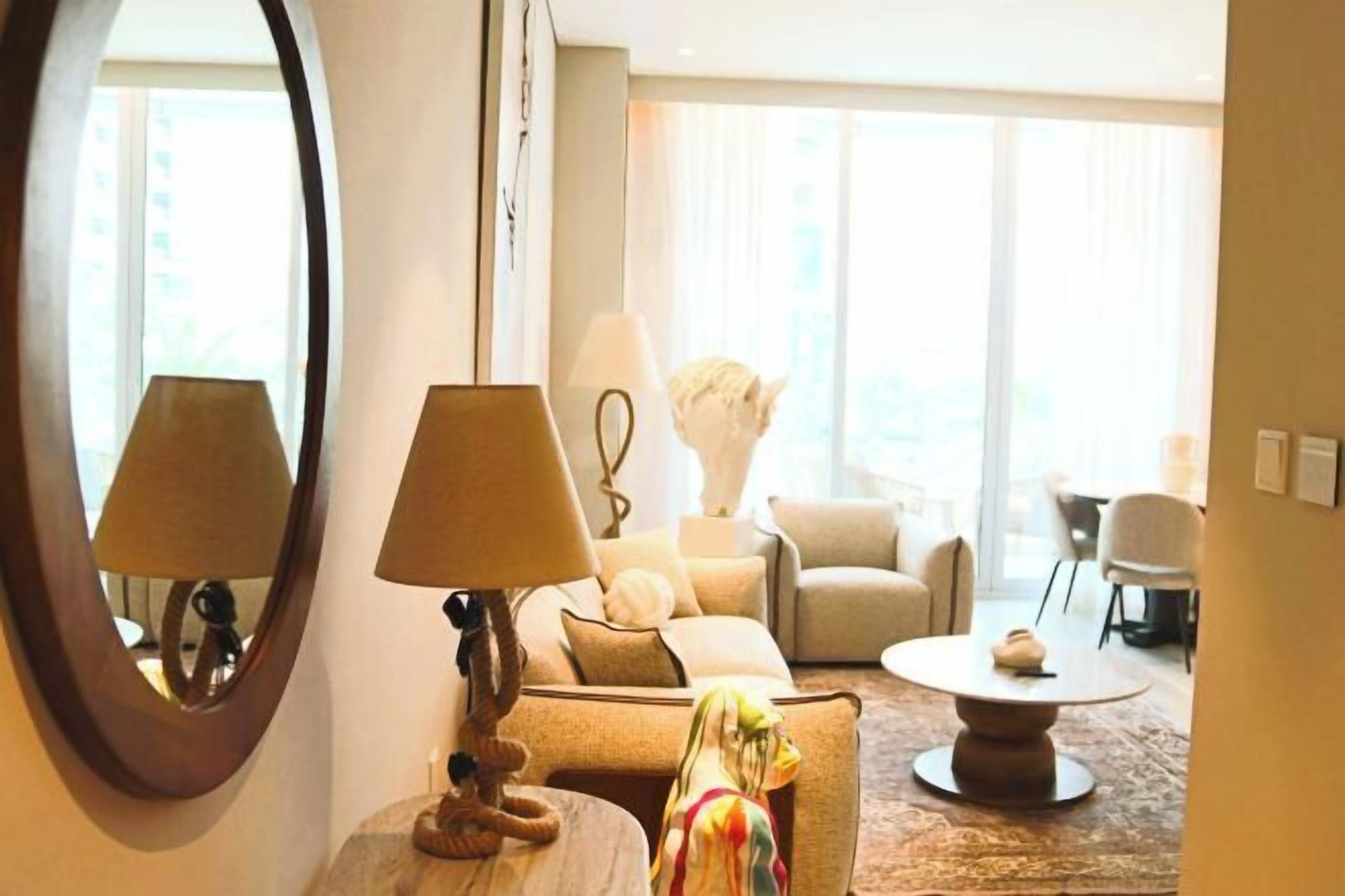 Uma sala de estar moderna com um sofá, uma mesa de centro redonda e um abajur. Há um espelho grande na parede e uma janela que proporciona iluminação natural.