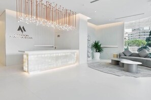 A modern reception area with a white reception desk, a unique hanging light fixture, and a grey sofa in a spacious room with a large window. - Casa Royale Vacation Home - Dubai Marina (Dubai)