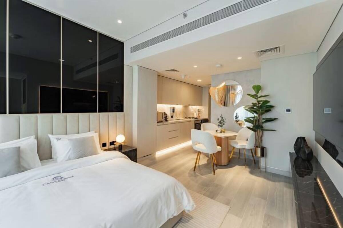 A modern hotel room with a large bed, a dining area, and a kitchen in the background.