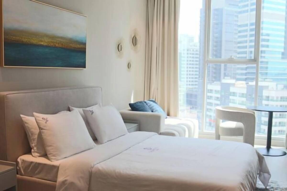 A hotel room with a bed, a sofa, a small table, and a large window with a city view.