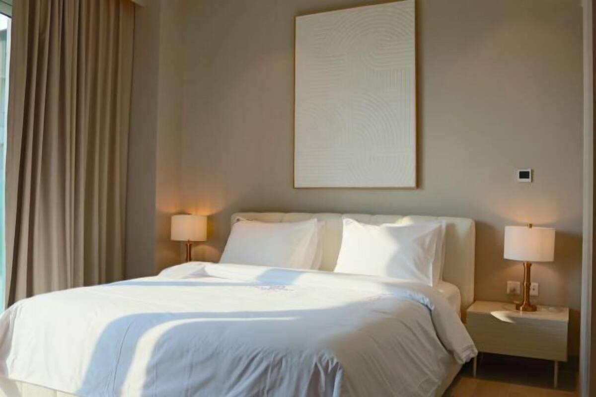 A hotel room with a bed, two bedside lamps, a large abstract painting on the wall, and a window with curtains.